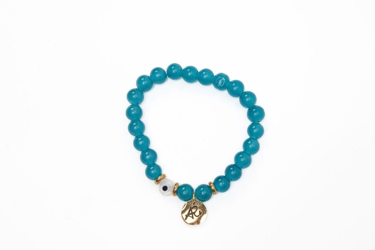 Teal beaded bracelet with a gold charm, gold accents, and a single white bead with a black dot on a white background.