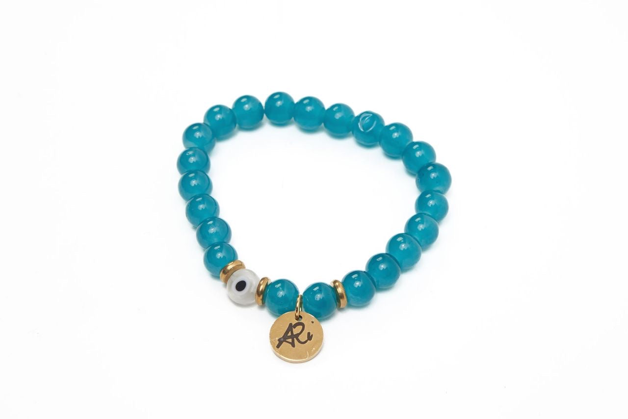 Teal beaded bracelet with a gold charm, gold accents, and a single white bead with a black dot on a white background.